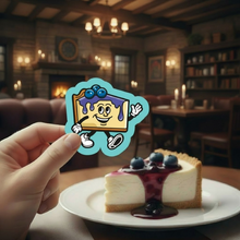 Load image into Gallery viewer, Hand holding a blueberry cheesecake cartoon character sticker over a slice of blueberry cheesecake in a cozy supper club dining room.
