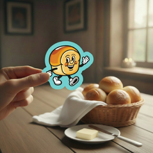 Load image into Gallery viewer, Hand holding a dinner roll cartoon character sticker over a basket of dinner rolls on a dining table.