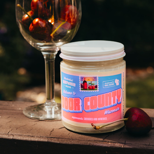 Door County soy candle in a 9 oz glass jar, from the Wanderlust Wisconsin Collection by Angela Rose Studios, shown outdoors on a wooden railing with a wine glass full of cherries beside it and a softly blurred, sun-dappled background.