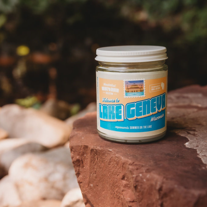 Lake Geneva soy candle in a 9 oz glass jar, from the Wanderlust Wisconsin Collection by Angela Rose Studios, shown outdoors on a rock with a blurred natural, outdoor-naturesque setting in the background.