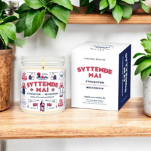 Load image into Gallery viewer, Syttende Mai in a 9 oz glass jar with packaging box, an exclusive seasonal release by Angela Rose Studios, shown on a wooden shelf next to an array of potted plants.
