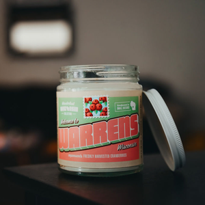 Warrens soy candle in a 9 oz glass jar, from the Wanderlust Wisconsin Collection by Angela Rose Studios, shown indoors on a dark wooden surface with a softly blurred, warm-toned background and the candle lid resting off to the side.